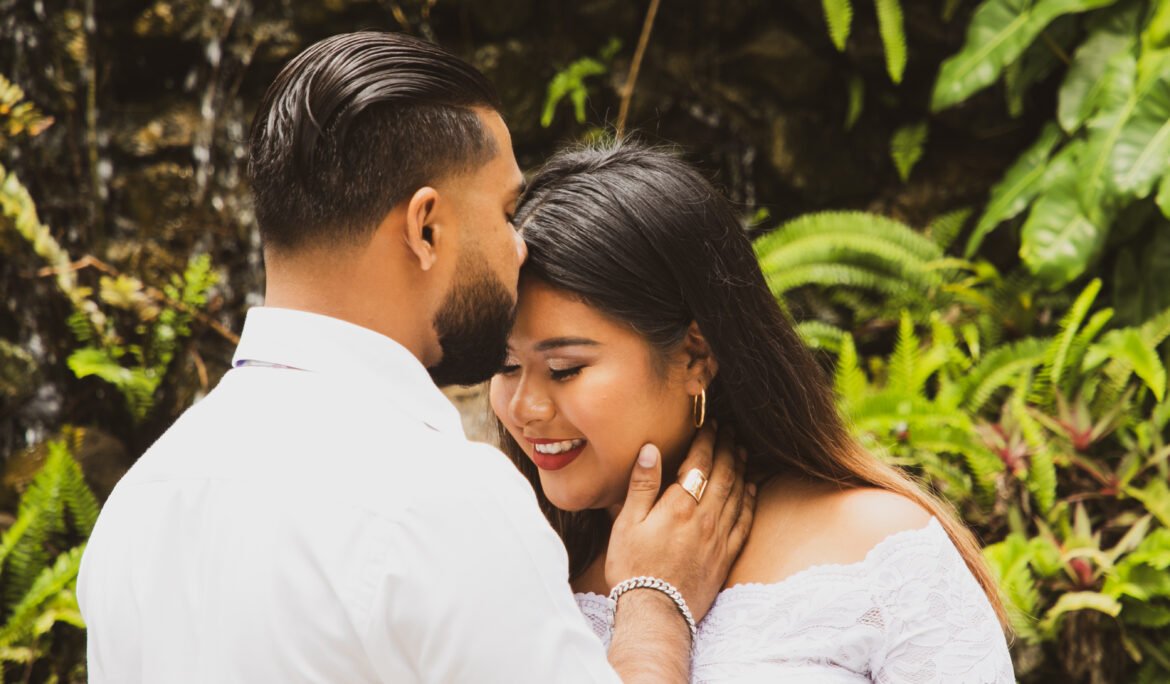 A married couple posing facing each other with the husband kissing her forehead